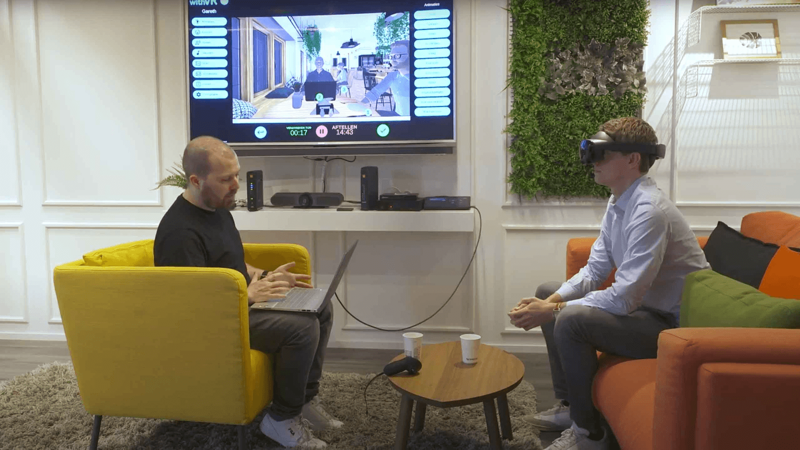 شاب يضع سمّاعة withVR بينما يدير الإكلينيكي جلسة التدرّب على التحدث من حاسوب محمول في الخلفية.
