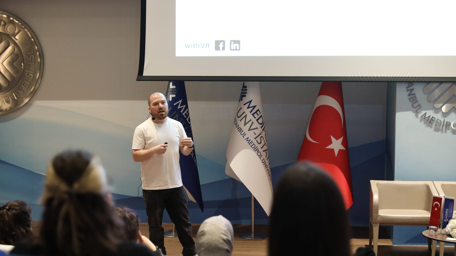 Gareth Walkom يلقي محاضرة ضيفية على المنصة في Medipol University، إسطنبول، وخلفه أعلام الجامعة والأعلام الوطنية